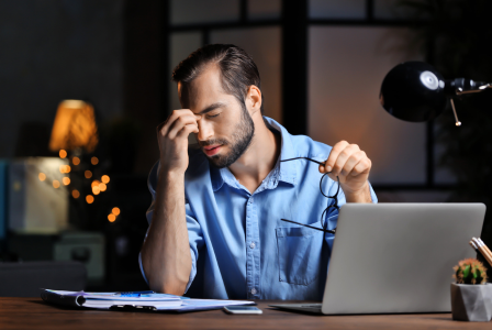 photo of a man working at a laptop and rubbing his eyes as if he is tired