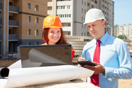 photo of lady and man at a building site with hard hats on working at a laptop and tablet